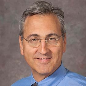 Headshot of a middle aged masculine-presenting person wearing round glasses and a blue button down shirt.