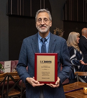 A smiling middle-aged man wearing a blue suit holds a gold plaque on a wood backing in his hands.