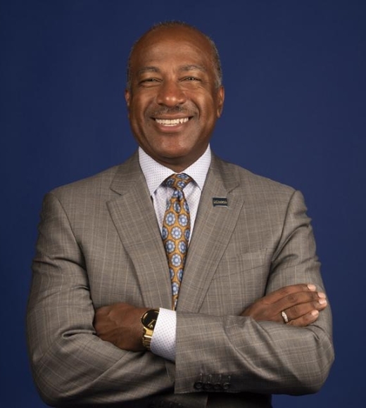 Chancellor Gary S. May wearing a gray suit and gold tie with light blue florals with arms folded in front of a dark blue background. He has a UC Davis lapel pin on the collar of his suit.