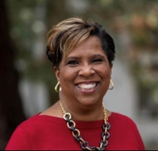 Frances Renty Williams with gold hoop earrings and a chunky gold and black chain necklace with a read knit sweater smiling outdoors.