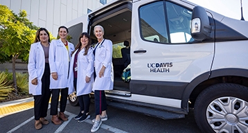 A group of four nurses wearing white coats and stethoscopes pose in front of a mobile health van.