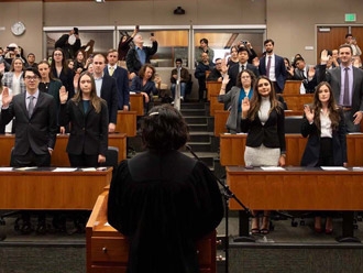 A judge leads a swearing in ceremony for law graduates after they pass the Bar exam. Sworn in attorneys wearing formal attire are standing with their right hand raised in the air.