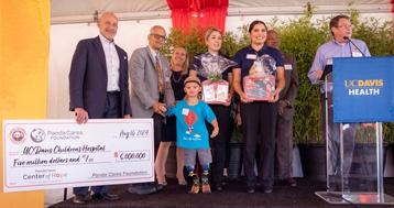 Administrators and a family pose onstage at a dedication ceremony with a podium positioned to the right. One person holds up a large check.