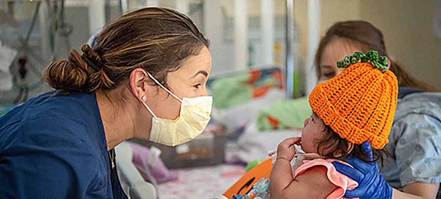 A physician wearing scrubs and a mask interacts with a child patient wearing an orange beanie in the shape of a pumpkin.