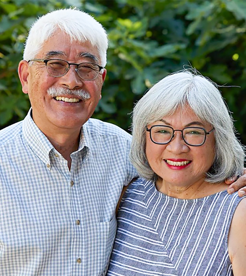 Sharon and Gary Takahashi pose in front of green foliage wearing glasses. Gary wears a blue and white gingham button-down shirt and Sharon wears a linen, striped, sleeveless top.