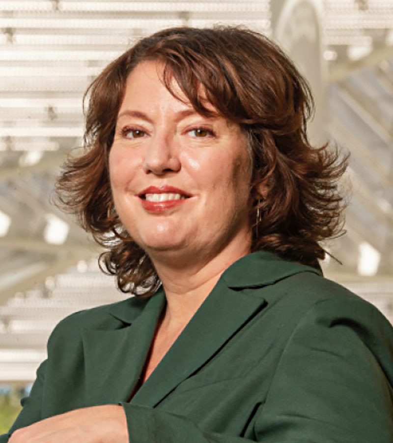 Rachel Teagle poses in a forest green blazer with the canopy structure of the Manetti Shrem Museum visible behind her.